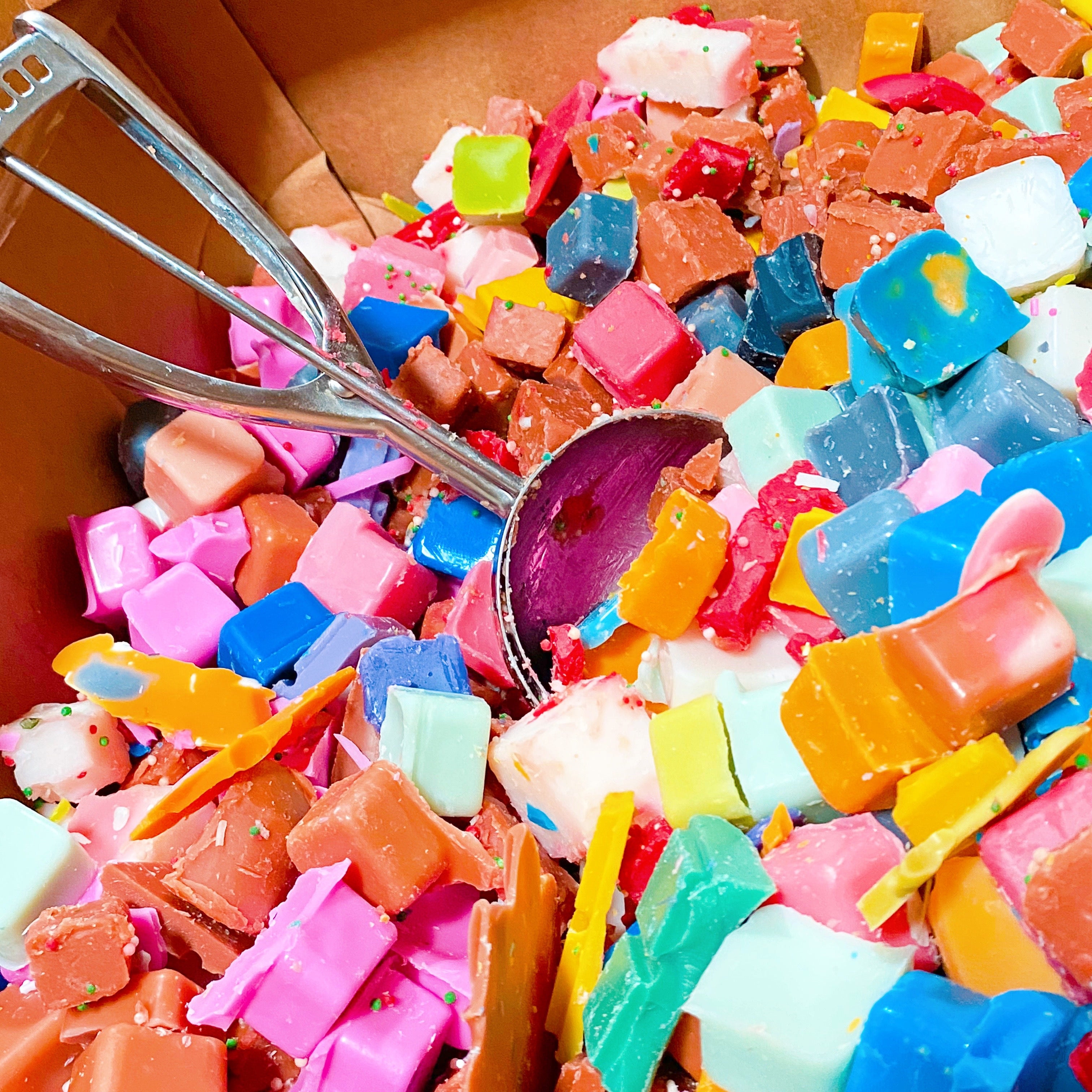 Colorful chunks of mystery wax in various bright shades with a metal scoop inside a cardboard box.